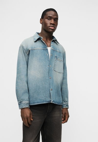 Νεαρός άνδρας που φοράει ανοιχτό μπλε πουκάμισο τζιν πάνω από λευκό εσώρουχο και σκούρο γκρι τζιν, μπροστά σε απλό φόντο.