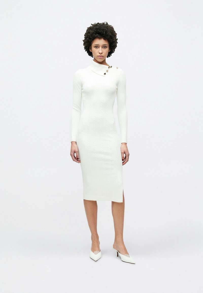 Λευκό πλεκτό φόρεμα με ριμπ ύφανση, μακριά μανίκια και ψηλό γιακά. Διαθέτει πλαϊνή σκίσιμο και διακοσμητικά χρυσά κουμπιά στον ώμο. Υποδήματα: λευκές γόβες.