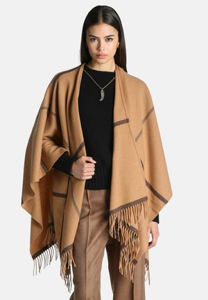 Donna con capelli lunghi e scuri che indossa un maglione nero, pantaloni marroni e uno scialle beige con frange e righe scure, in piedi davanti a uno sfondo neutro.