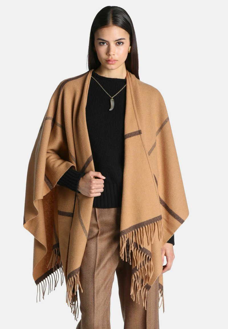 Femme aux longs cheveux foncés portant un pull noir, un pantalon marron et un châle beige à franges avec des rayures foncées, debout devant un fond uni.