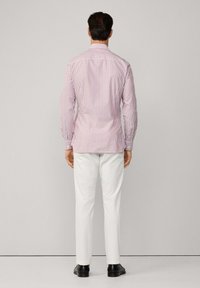 Košile s knoflíky v červenobílých vertikálních pruzích, bavlněný materiál, dlouhé rukávy, přizpůsobený střih, nošena s bílými kalhotami, zobrazeno ze zadní strany.