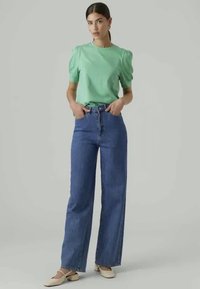 Zöld, puffos ujjú felső, magas derekú, bőszárú kék farmernadrággal párosítva. A ruházat laza szabású és minimalista dizájnú.