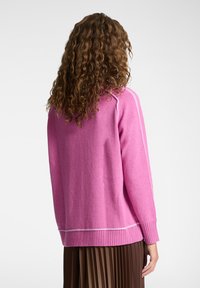 Maglione rosa con orlo e polsini a coste, caratterizzato da una texture liscia e dettagli di cucitura a contrasto lungo le spalle. Gonna marrone plissettata.
