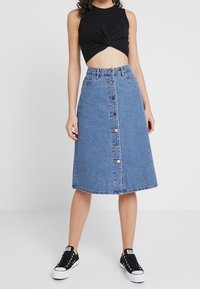 Falda de mezclilla en línea A, azul claro, hasta la rodilla, con cierre de botones en el frente y bolsillos traseros. Combinada con un top corto negro y zapatillas.