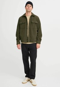 Chaqueta de forro polar verde con cierre de botones, dos bolsillos en el pecho, usada sobre una camisa beige, combinada con pantalones negros y zapatillas blancas.