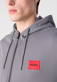 Siva dukserica s patent zatvaračem i kapuljačom s elastikom. Ima crveni pravokutni uložak s natpisom "HUGO" u crnoj boji. Tekstura tkanine izgleda mekano.