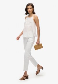 Witte mouwloze top met kantdetails, gecombineerd met witte slim-fit broek, bruine haksandalen en een kleine geweven tas.