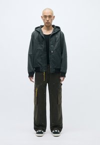 Μαύρο φούτερ με κουκούλα και φερμουάρ, διαθέτει μπροστινές τσέπες και κίτρινες κορδόνες. Ελαιόχρονες cargo παντελόνες με πολλές τσέπες και φερμουάρ. Μαύρα αθλητικά παπούτσια.