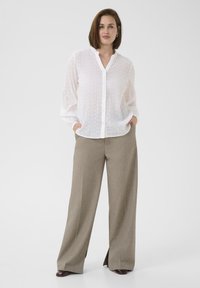 Witte, lichtgewicht overhem met een gestructureerd patroon, gecombineerd met losse, beige hoge taille broek. Model staat met handen in de zakken.