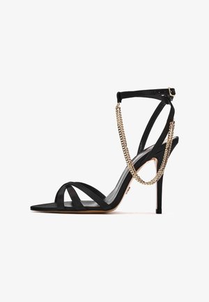 Sandale noire à talon haut avec des lanières fines, une boucle à la cheville et une chaîne décorative dorée drapée autour de la lanière de la cheville.