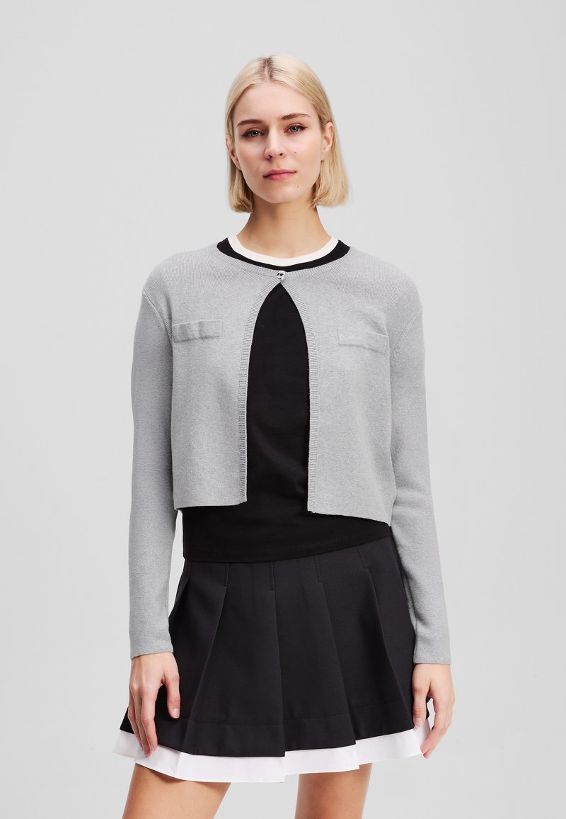 Cardigan grigio a maniche lunghe con scollo rotondo, chiusura con bottoni e un taglio corto. Presenta due tasche frontali e una texture a coste. Top nero e gonna plissettata.