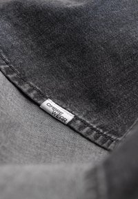 Zwarte denimstof heeft een gladde textuur met contrasterende stiksels. Een wit label met "OMBRE WEAR" is zichtbaar langs de naad.