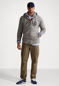 Γκρι φούτερ με φερμουάρ και το κείμενο «Polo», φορεμένο πάνω από μπλε πουκάμισο. Μπεζ παντελόνι cargo και μπλε αθλητικά παπούτσια ολοκληρώνουν τη στολή.