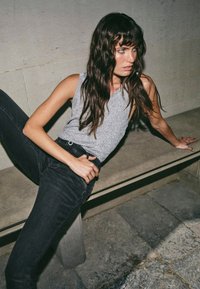 Sivi top bez rukava s teksturiranim završetkom, uparen s uskim crnim trapericama. Osoba pozira na kamenom klupcu uz svijetlu pozadinu.