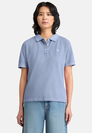 Femme aux cheveux noirs mi-longs portant un polo à manches courtes bleu clair et un jean en denim bleu clair, debout devant un fond blanc.