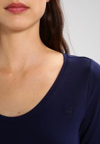 Mörkblå figursydd skjorta med djup V-ringning, slät textur och en subtil präglad logotyp på bröstet.