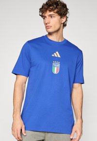 Tricou albastru din bumbac, cu guler rotund, mâneci scurte, logo Adidas auriu și o emblemă Italia cu patru stele deasupra.