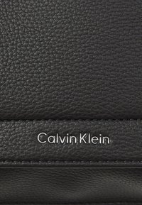 Black textured leather with a subtle grain. Features a silver "Calvin Klein" logo and stitched accents on the surface.
