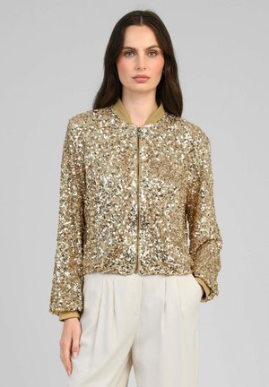 Femme portant une veste bomber à sequins dorés avec fermeture éclair, poignets et col beige, associée à un pantalon crème taille haute.
