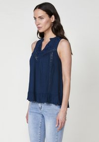 Top senza maniche color navy con ricami floreali sul collo, texture plissettata, orlo arricciato e bottoni decorativi lungo la parte anteriore.