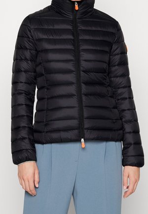 Schwarze gesteppte Jacke mit Reißverschluss und orangefarbenen Zippern, getragen zu hellblauen Hosen, gezeigt von den Schultern bis zur Taille.