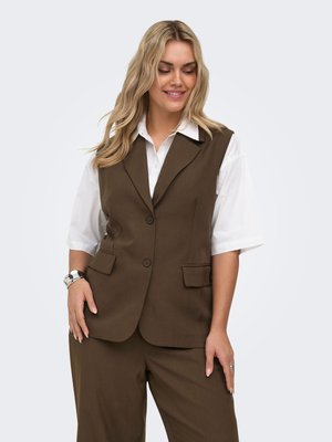 Femme aux longs cheveux blonds portant un blazer marron sans manches sur une chemise blanche et un pantalon marron assorti, souriante et posant avec une main dans la poche.