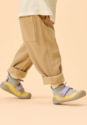 Child wearing beige rolled-up pants and knitted shoes with yellow and purple accents walking on light background.