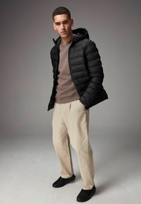Sort polstret jakke med hætte, brun sweater, beige bukser og sorte sneakers. Lige pasform og glatte teksturer. Indendørs studieindstilling.