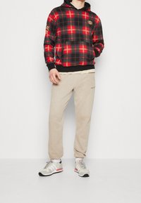 Felpa con cappuccio a quadri nei colori rosso, nero e oro, con tasca frontale, abbinata a pantaloni della tuta beige con un logo del brand discreto, indossati con sneaker.