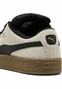 A Puma sportcipő bézs velúr felsőrésszel rendelkezik, fekete textúrázott swooshtal, párnázott nyakkal és barna gumitalppal, amelyen lézeres logó található.