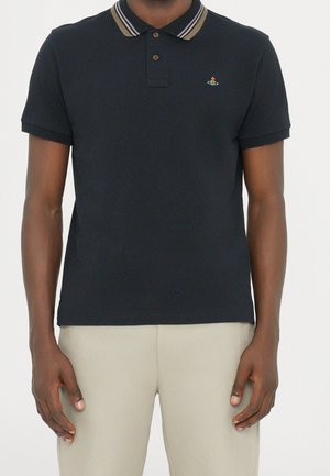 Man draagt een zwart poloshirt met korte mouwen, gestreepte kraag en klein kleurrijk logo op de linkerborst, gecombineerd met beige broek.
