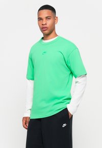 Tricou verde din bumbac, cu guler rotund și mâneci scurte, având un mic logo Nike în nuanța teal pe piept, purtat peste o cămașă albă cu mânecă lungă.