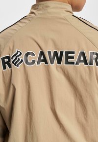 Svetlá bunda s vysokým golierom a čiernymi pruhmi. Na chrbte sa nachádza veľký, výrazný nápis "ROCAWEAR" v čiernej a bielej farbe.