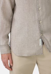 Camicia a maniche lunghe marrone chiaro realizzata in tessuto texturizzato, con bottoni, orlo curvo e un'etichetta di stoffa visibile vicino al fondo.