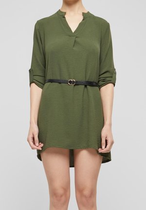 Personne portant une courte robe vert olive avec des manches retroussées et une fine ceinture noire avec une boucle double anneau dorée.