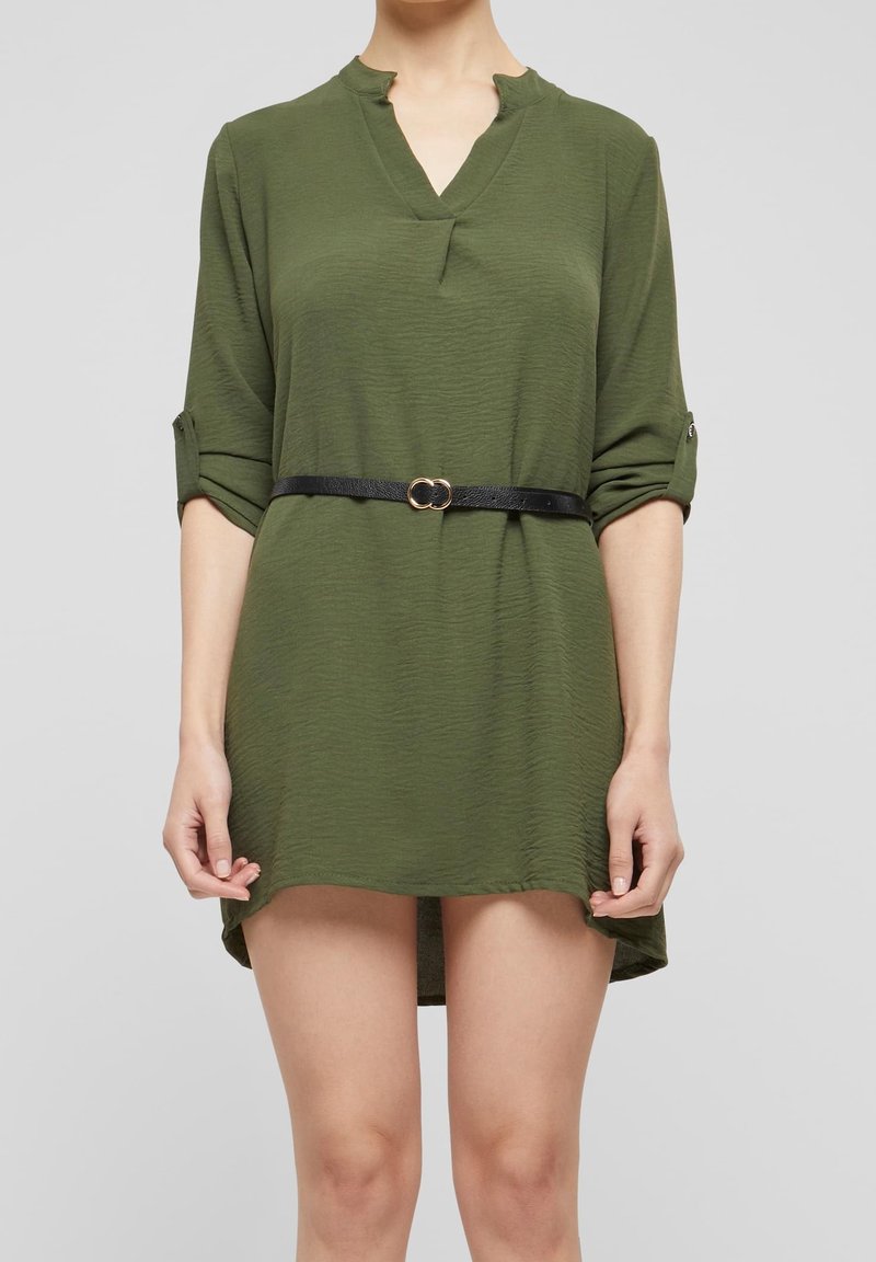 Personne portant une courte robe vert olive avec des manches retroussées et une fine ceinture noire avec une boucle double anneau dorée.