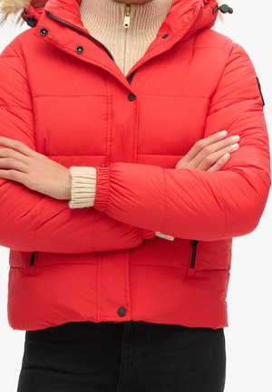 Osoba ubrana w jasnoczerwoną puchową kurtkę z czarnymi guzikami i kieszeniami na zamek, z rękami skrzyżowanymi na beżowym dzianinowym swetrze.