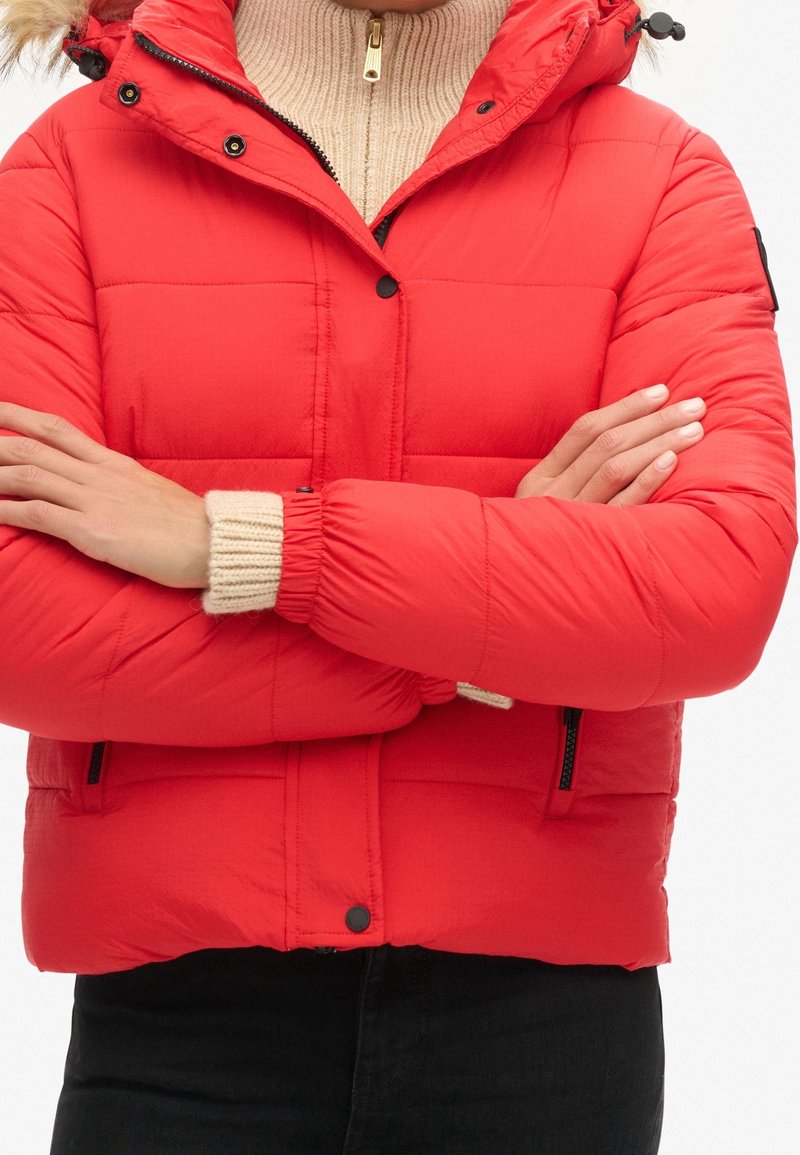Personne portant une doudoune rouge vif avec des boutons noirs et des poches zippées, les bras croisés sur un pull en maille beige.
