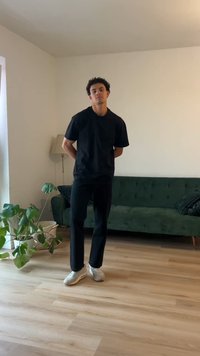 Ung mann med krøllete hår står i en minimalistisk stue iført svart t-skjorte, svarte bukser og hvite joggesko ved en grønn sofa og planter.