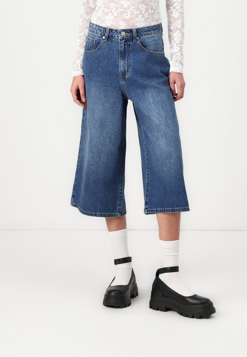 Culotte din denim de culoare albastru mediu, cu talie înaltă, croială lejeră și umeri brut. Asortate cu pantofi cu platformă negri și șosete albe.