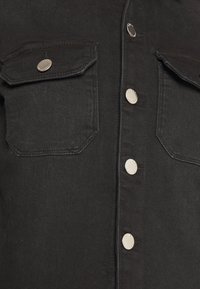 Chemise en denim noir avec deux poches avant et fermetures à boutons en métal, possédant une surface propre et texturée ainsi qu'une quincaillerie argentée contrastante.