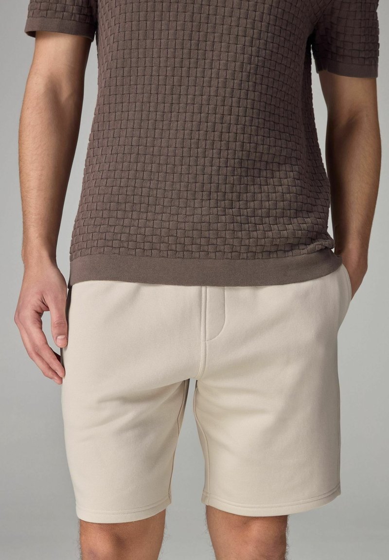 Homme portant un short beige et une chemise marron texturée à manches courtes avec un motif à carreaux, debout les mains le long du corps.