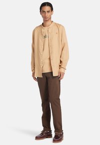 Camicia button-down beige chiaro sopra una maglietta color sabbia, abbinata a pantaloni marroni e scarpe rosse scure con accenti e lacci beige.