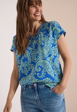 Femme souriante aux longs cheveux bruns portant une chemise bleu vif à motifs paisley verts et un jean en denim bleu, une main dans la poche.