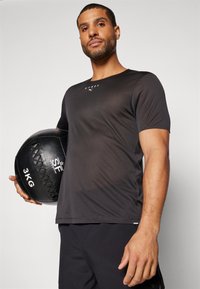 Svart trenings t-skjorte med korte ermer, med et teksturert mønster og en liten hvit logo. Modellen holder en svart 3 kg medisinball.