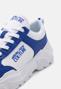 White and blue sneaker with mesh upper, leather accents, thick sole, and "Versace Jeans Couture" logo on the side.