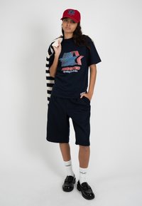 Joven mujer con gorra roja, camiseta azul marino con diseño gráfico, pantalones cortos a juego, calcetines blancos, mocasines negros, sosteniendo un suéter de rayas sobre el hombro sobre un fondo liso.