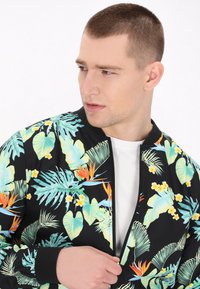 Fekete bomber dzseki trópusi mintával, zöld levelekkel, narancssárga virágokkal és sárga részletekkel. Simító anyagból készült, cipzáros záródással.