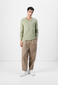 Lys grønn v-hals genser med lange ermer, kombinert med beige vide bukser og hvite sneakers. Minimalistisk design med subtle logo detaljer.