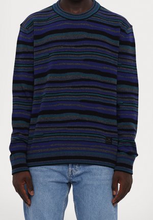 Homme portant un pull à manches longues bleu, noir et vert à rayures horizontales avec un jean bleu clair, debout sur fond blanc.